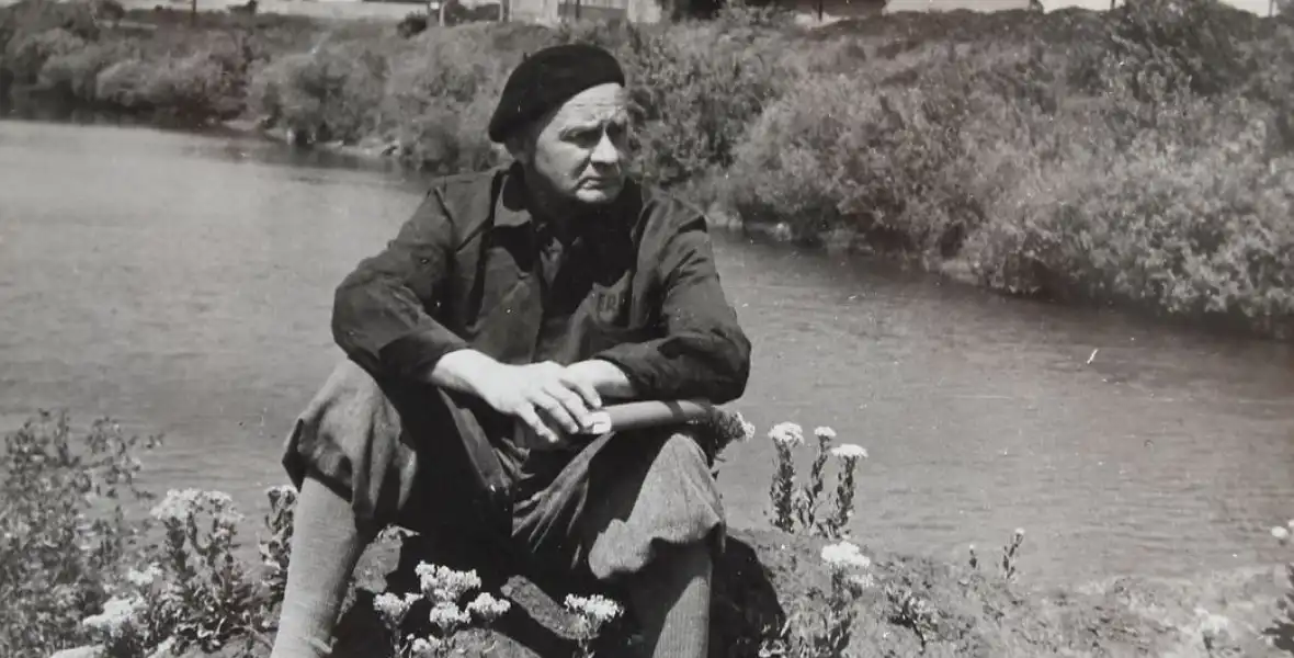 Hamvas Béla író a folyóparton ül, kezében könyv, a fején baszk sapka, fekete fehér kép